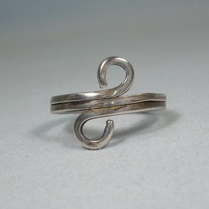 May include: A silver-toned ring with a unique, open design. The ring features two curved, swirling elements that meet in the center, creating an eye-catching, abstract shape. The band is a simple, double-layered design. The ring is set against a light background.