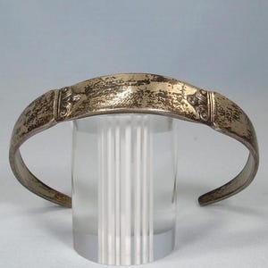 May include: A silver-toned cuff bracelet with a textured, aged appearance. The bracelet features decorative elements on each end and is displayed on a clear, cylindrical stand. The design suggests a vintage or antique style.