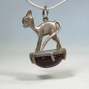 May include: A silver-toned cat pendant with a stylized design. The cat stands atop a dark red, half-circle base. The pendant is suspended from a silver chain, creating a vintage or antique aesthetic. The cat's tail curves gracefully.