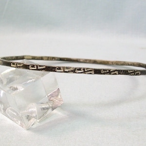 May include: A silver-toned bangle bracelet with a textured, rectangular pattern. The bracelet is displayed on a clear, geometric stand against a white background. The bangle's design features a series of small, raised rectangular shapes.