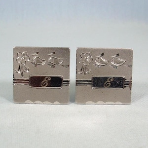 May include: A pair of square silver cufflinks. Each cufflink features a floral and leaf design at the top, a horizontal stripe, and a rectangular section with the letter "E". The bottom edge has a scalloped design.