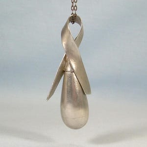 May include: A silver-colored pendant with a teardrop-shaped body and a ribbon-like design at the top. The pendant hangs from a delicate chain. The metal has a brushed finish, giving it a subtle sheen. The overall design is minimalist and elegant.