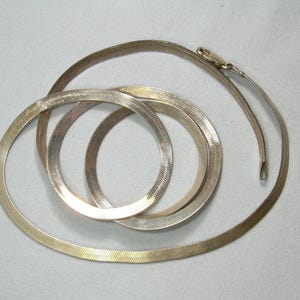 May include: A collection of gold and silver-toned herringbone necklaces and bracelets. The jewelry is arranged in a circular pattern, with a clasp visible on one necklace. The pieces have a smooth, reflective surface.