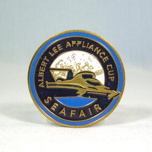 May include: A round pin with a blue and gold border. The pin features the text "ALBERT LEE APPLIANCE CUP SEAFAR" in gold lettering. A gold boat is in the center of the pin, with a white background.