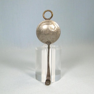 May include: A silver spoon with a circular coin-like head featuring a profile of a Native American and the word "LIBERTY". The spoon has a long, slender handle and a small ring at the top. The spoon is displayed on a clear, cylindrical stand.