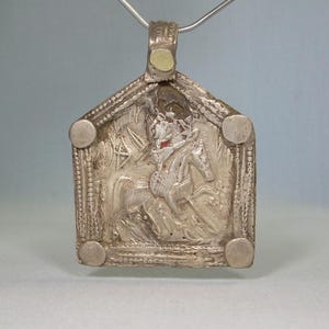 May include: A silver pendant with a rectangular frame and a detailed relief of a figure on horseback. The pendant has a loop at the top for a chain and four circular embellishments on the corners.