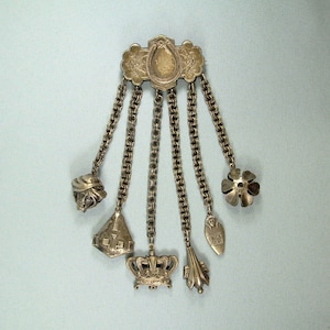Può includere: Un braccialetto di charme in argento con una parte superiore decorativa e cinque ciondoli pendenti. I ciondoli includono una corona, un fiore, una campana, un cappello e uno scudo.