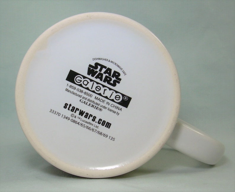 May include: White ceramic mug with a black Star Wars logo and Galerie branding. The mug is dishwasher and microwave safe. The bottom of the mug has the text "starwars.com" and a phone number.