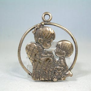 Puede incluir: Un colgante plateado que representa a una madre y un niño dentro de un marco circular. La madre está sentada en una silla con respaldo acolchado, y el niño está de pie a su lado. Un pequeño osito de peluche está a sus pies. El colgante tiene un lazo en la parte superior para colgar.