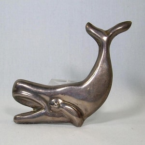 May include: A silver whale sculpture with a smooth, polished finish. The whale is depicted in a stylized, abstract form with a curved body and a long, pointed tail.