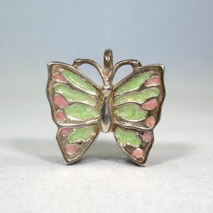 Puede incluir: Un colgante de mariposa plateado con detalles de esmalte verde y rosa. La mariposa tiene un diseño de alas detallado y un lazo para una cadena. El colgante es un accesorio de joyería.