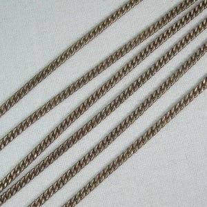 Peut inclure: Gros plan de plusieurs chaînes gourmette dorées disposées en diagonale sur un fond blanc. Les chaînes ont un design classique à maillons imbriqués et le métal a un aspect légèrement vieilli.