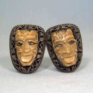 May include: A pair of vintage cufflinks featuring carved faces with smiling expressions. The faces are a light tan color, set within ornate, oval-shaped silver-toned frames. The cufflinks are displayed on a white surface.