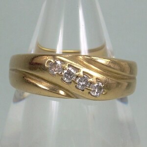 May include: A gold-colored ring with four small, clear gemstones set diagonally across the top. The ring has a textured, ridged design on the band. The ring is displayed on a clear, cone-shaped stand.