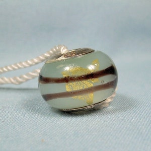 Puede incluir: Una cuenta de charm de plata con un diseño de vidrio rayado azul claro y marrón. El vidrio tiene un patrón de hoja de oro y un núcleo de plata.