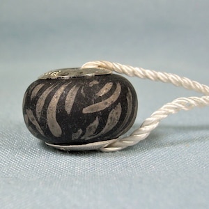Peut inclure: Un charme de perle noir et argent avec un motif en spirale. La perle est attachée à un cordon blanc.