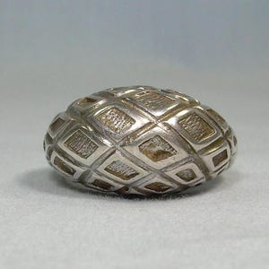 May include: A silver-colored, oval-shaped button with a repeating diamond pattern. The button has a textured surface and a slightly rounded form. The design is symmetrical, with each diamond shape clearly defined. The button is likely for clothing or crafts.