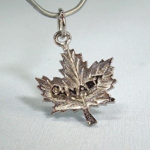 May include: Silver-toned maple leaf pendant with the word "CANADA" inscribed across the center. The pendant is attached to a small silver loop, suitable for a necklace or bracelet. The leaf design is detailed and textured.