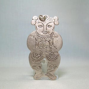 Puede incluir: Pendiente de plata con un diseño en relieve detallado de una persona sosteniendo un objeto redondo. El pendiente tiene un bucle en la parte superior para colgar.