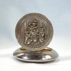 May include: A silver-toned, hinged compact with a detailed relief scene on the lid. The scene depicts figures in a religious setting. The compact is open, revealing a small, oval base.