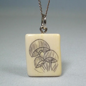May include: A cream-colored, square pendant necklace with a silver chain. The pendant features a detailed black ink drawing of several mushrooms. The necklace is a simple, elegant accessory.
