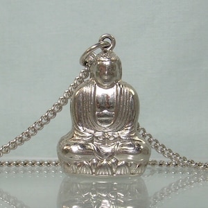 May include: Silver pendant necklace featuring a detailed sculpture of a seated Buddha. The chain is silver and has a delicate link design.