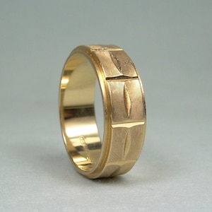 Könnte beinhalten: Ein goldfarbener Ring mit strukturierter Oberfläche und einem sich wiederholenden Muster aus vertikalen und horizontalen Linien. Der Ring hat ein leicht abgerundetes Profil und eine polierte Oberfläche. Das Design ist symmetrisch.