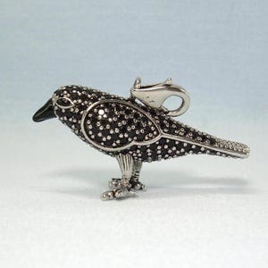 Puede incluir: Un colgante de pájaro en tono plateado con un pico negro y un cuerpo cubierto de pequeñas piedras oscuras y brillantes. El pájaro se muestra de perfil, de pie sobre dos patas, con un cierre de mosquetón de metal en la parte superior del lomo.