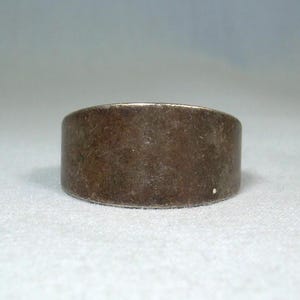 May include: A wide, silver-toned ring with a brushed metal appearance. The ring has a simple, band-style design and a slightly worn look. The ring is displayed on a white surface.