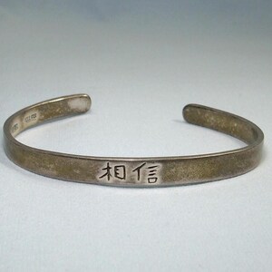 May include: A silver-toned cuff bracelet with the Chinese characters for "believe" engraved on the front. The open-ended bracelet has a slightly tarnished appearance, suggesting age or wear. The bracelet is displayed on a light-colored surface.