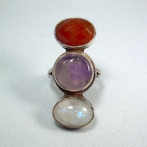 Könnte beinhalten: Ein Silberring mit drei Edelsteinen: einem facettierten orangefarbenen Karneol, einem runden lila Amethyst und einem ovalen Mondstein. Die Edelsteine sind in einzelnen Fassungen gefasst, wodurch ein gestapeltes Design entsteht.