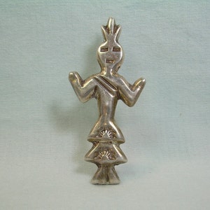 May include: A silver charm depicting a stylized Native American figure with a headdress and a tiered skirt. The figure is standing with arms outstretched.