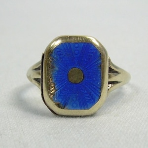 May include: A gold-toned ring with a rectangular blue enamel face. The blue enamel features a central dark circle and radiating patterns. The ring has a classic design with a simple band.
