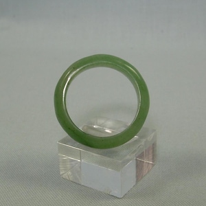 Puede incluir: Un anillo de jade verde, mostrado en un soporte de acrílico transparente. El anillo es una banda simple con una superficie lisa y pulida.