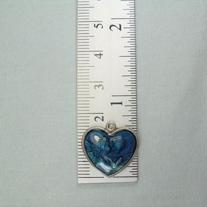 May include: A silver heart-shaped charm with a blue image of two angels inside. The charm is about 1 inch in diameter.