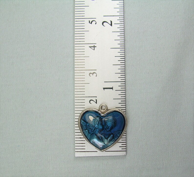 May include: A silver heart-shaped charm with a blue image of two angels inside. The charm is about 1 inch in diameter.