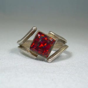 May include: A silver ring with a red and iridescent opal gemstone set in a geometric design.