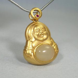May include: A gold-colored pendant featuring a smiling Buddha figure with a light-colored stone in the center. A small red gemstone is set in the loop at the top. The pendant hangs from a silver-colored chain.