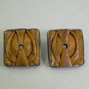 May include: A pair of square-shaped earrings with a carved bone design. The bone is a light brown color and the earrings are set in silver.
