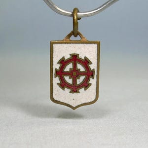 May include: A small, vintage pendant with a white background and a red and gold design. The pendant is shield-shaped with a gold border and a red gear-like symbol in the center. It hangs from a silver chain.