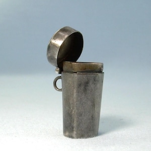 Könnte beinhalten: Ein kleiner, verwitterter silberfarbener Metallbehälter mit einem geöffneten Klappdeckel. Das Gefäß hat einen konisch zulaufenden, rechteckigen Körper und eine kleine kreisförmige Öse an einer Seite, mit einer stumpfen, gealterten Metalloberfläche.