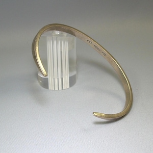 May include: A gold-toned metal bangle bracelet with a simple, curved design. The bracelet has a small inscription on the inside: "#181 PCD001 0729".