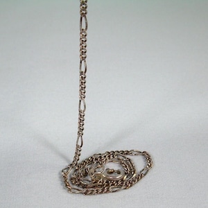 May include: A silver-colored Figaro chain necklace. The chain is laid out on a white surface, with a portion of the chain standing upright. The chain has a repeating pattern of three small links followed by one elongated link.