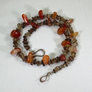 以下が含まれることがあります： 銀色の留め具が付いた、茶色とオレンジ色の宝石のネックレス。ネックレスは、小さく不規則な形の石で作られています。