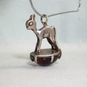 Puede incluir: Pendiente de plata con un gato estilizado de pie sobre una piedra preciosa redonda roja. El pendiente está unido a una cadena de plata.