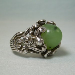 May include: A silver ring featuring a green jade stone set within an intricate floral design.