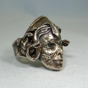 May include: A silver ring with a detailed, sculpted face with a sinister expression. The ring has a textured finish and a design that includes a hat and coiled elements around the face. The ring is likely a piece of jewellery.