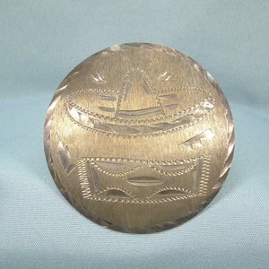 Puede incluir: Broche de tono plateado con un diseño de sombrero. La broche es redonda y tiene una superficie texturizada.