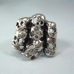 Puede incluir: Un anillo plateado con un grupo de diseños de calaveras. El anillo tiene una apariencia detallada y tridimensional con múltiples calaveras dispuestas juntas. Las calaveras tienen cuencas de los ojos abiertas y rasgos ahuecados.