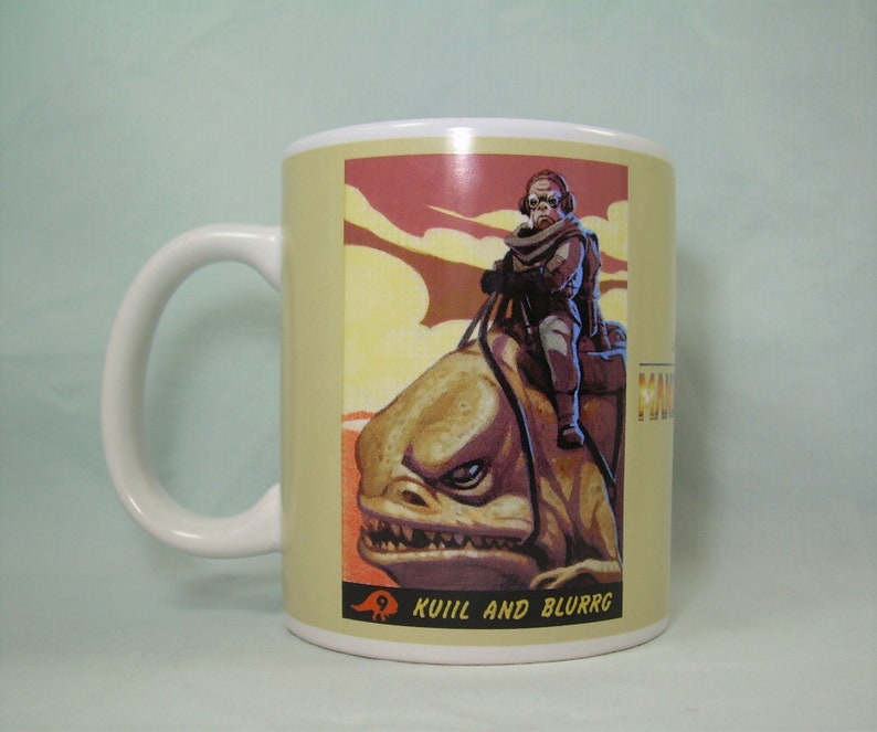Puede incluir: Una taza de cer&aacute;mica blanca con una ilustraci&oacute;n colorida de un personaje montando una criatura. La ilustraci&oacute;n es de estilo retro con una gama de colores rojo, naranja y amarillo. El texto "Kuiil and Blurrgg" est&aacute; impreso en la parte inferior de la ilustraci&oacute;n.